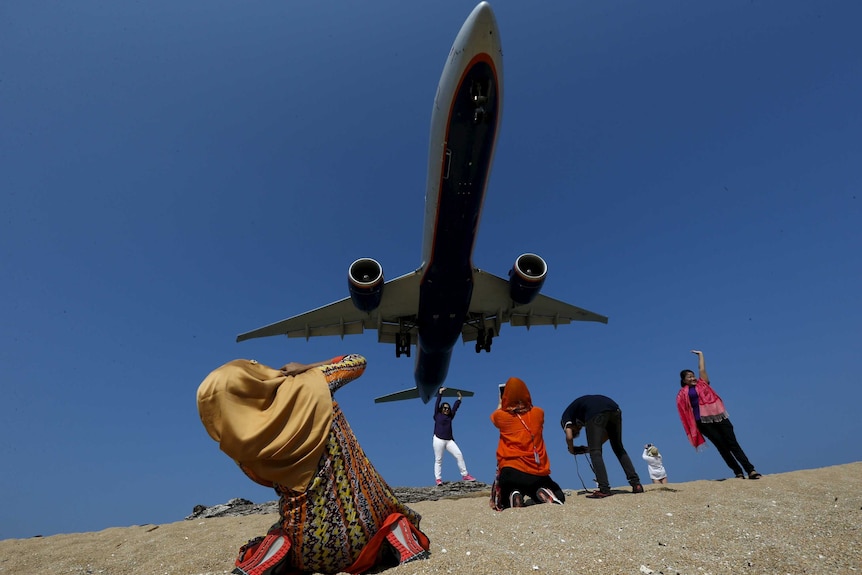 People crouch and pose on the sand taking photos and posing as a plane looms in the blue sky overhead