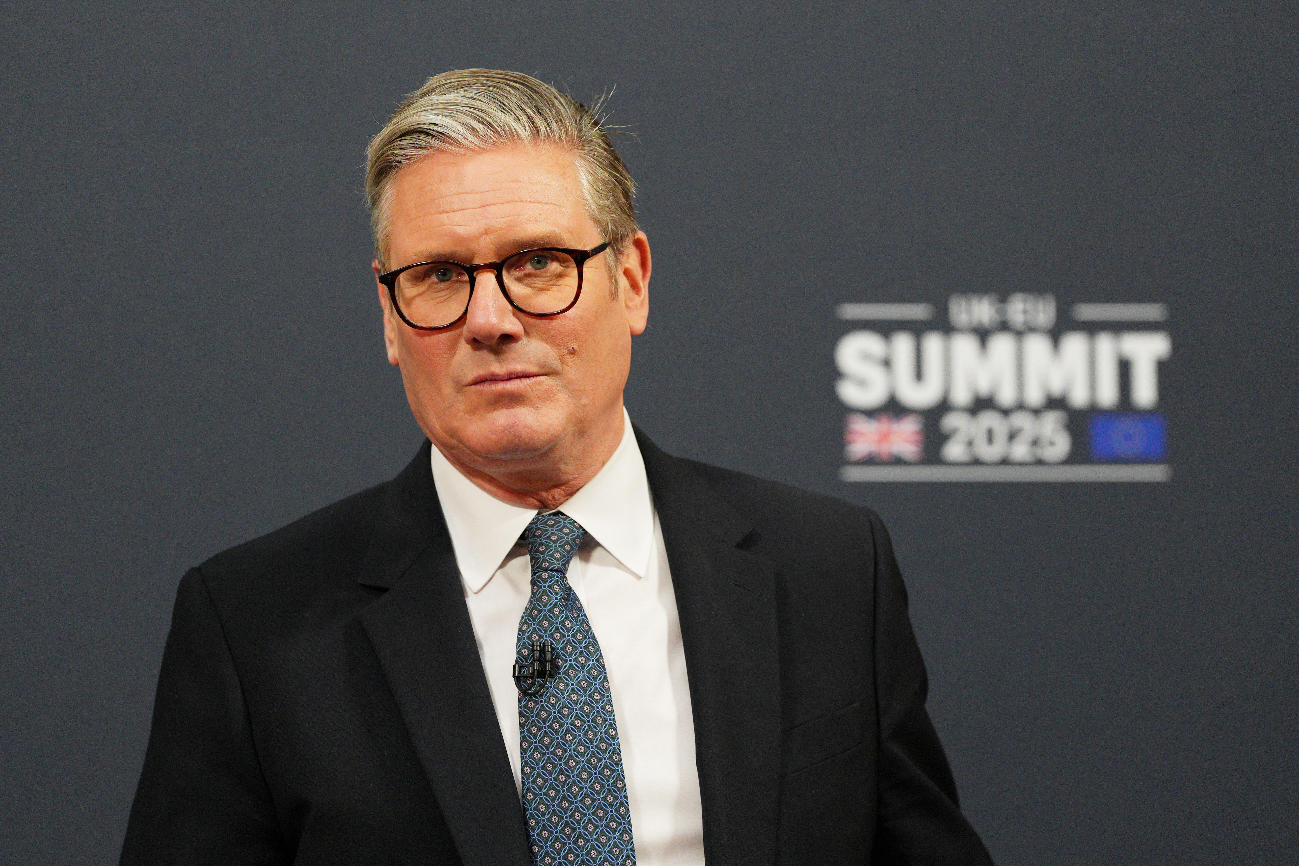 Keir Starmer wearing a suit and black-rimmed glasses speaking at a press conference in front of a grey background.