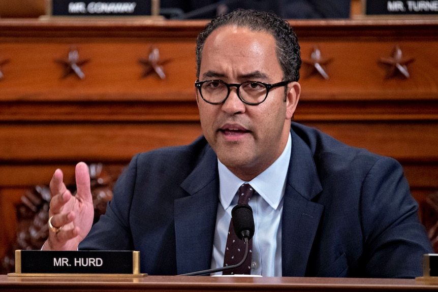 A middle-aged black man in a suit with glasses speaks while seated in front of an intricate wooden backdrop.