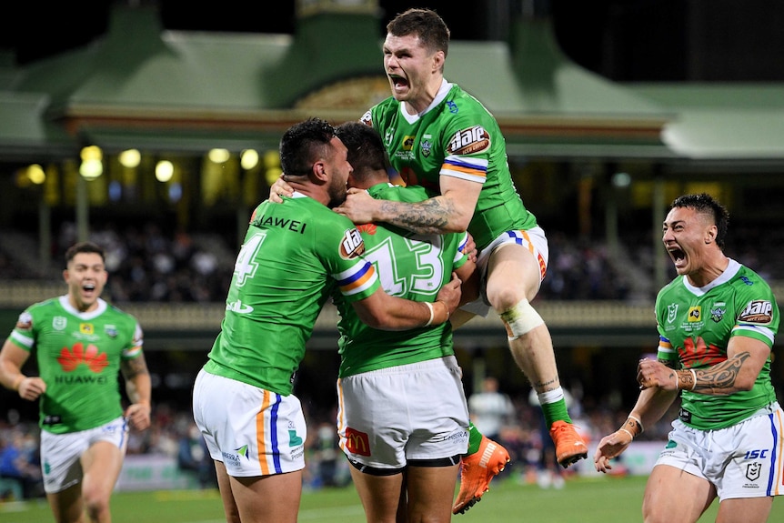 A Canberra Raiders NRL player jumps in the air as he embraces two teammates after a try was scored against the Sydney Roosters.