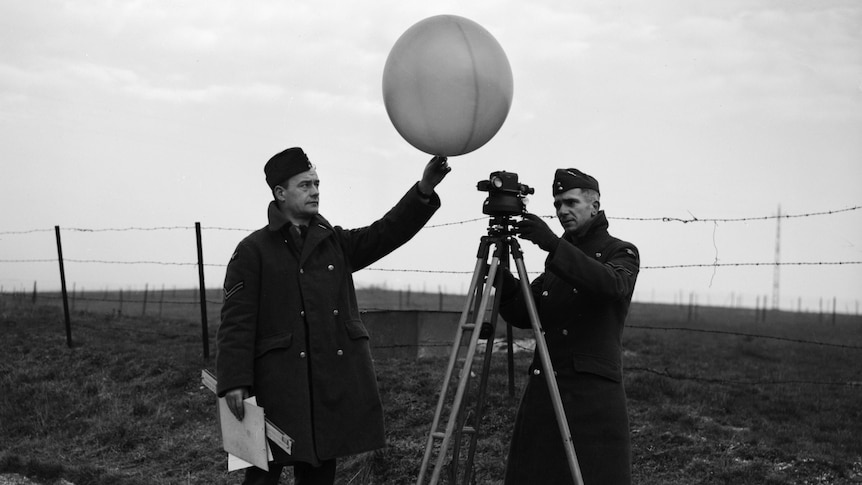 A black and white photo of two men in air force uniforms - one holding a weather balloon and one looking through an instrument.