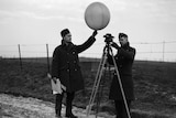 A black and white photo of two men in air force uniforms - one holding a weather balloon and one looking through an instrument.