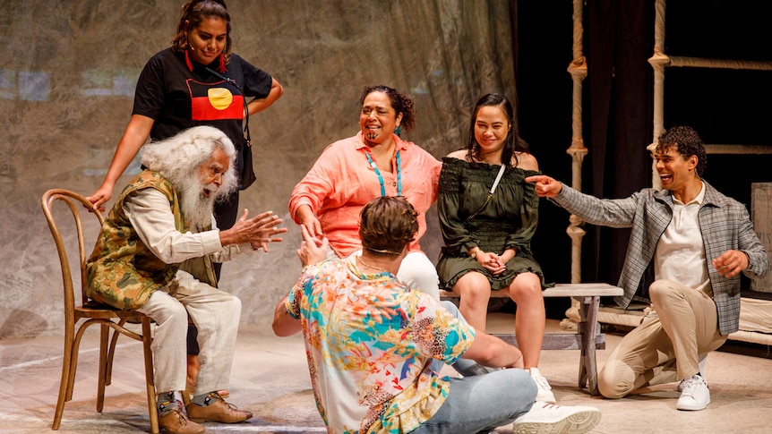 Stage shot showing group of six people sitting in a loose circle (and one young woman standing) as old man tells them story.