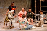 Stage shot showing group of six people sitting in a loose circle (and one young woman standing) as old man tells them story.