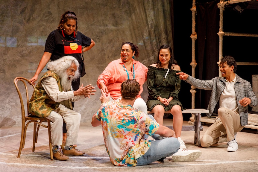 Stage shot showing group of six people sitting in a loose circle (and one young woman standing) as old man tells them story.