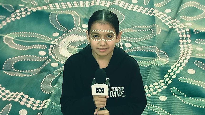 A girl with indigenous face paint sits in front of an indigenous art based fabric holding an ABC microphone.