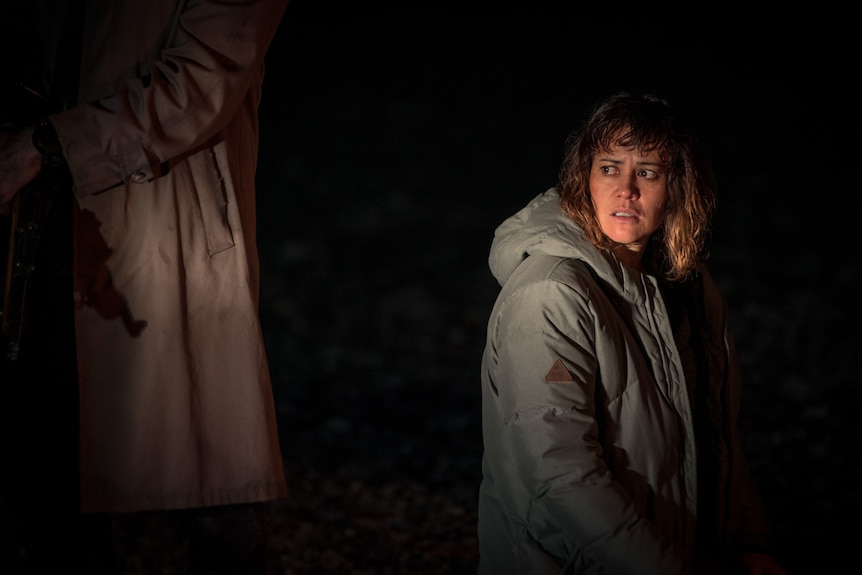 A Maori woman in her 40s looks afraid. She is standing in the dark, looking behind her at an unseen threat. 