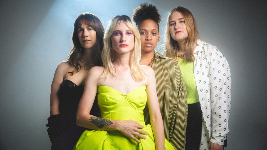 Four women stand in a spotlight in various outfits in the same acid green, white and black colour palette.