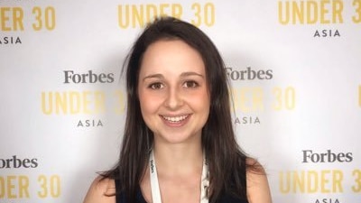 A young woman with brown hair smiling in front of a 'Forbes under 30' media banner