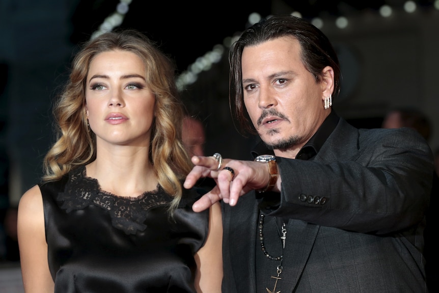 Amber Heard in a black dress with lace detail gazes off camera, next to Johnny Depp in a black suit, pointing