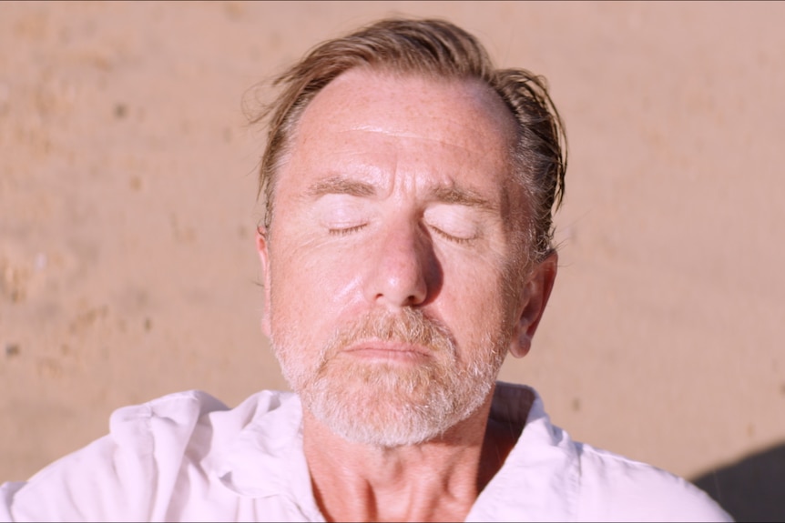 White man with graying beard and short mousey hair stands on beach with eyes closed and face tilted towards the sun.