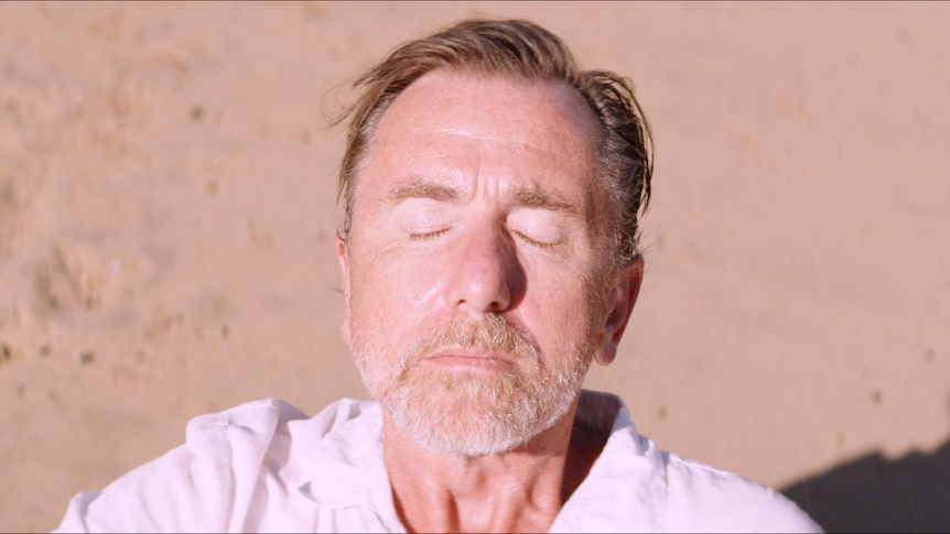 White man with greying beard and short mousey hair stands on beach with eyes closed and face tilted towards the sun.