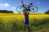 A cyclist with a prosthetic leg stands with a bike lifted above his head in a field of canola.