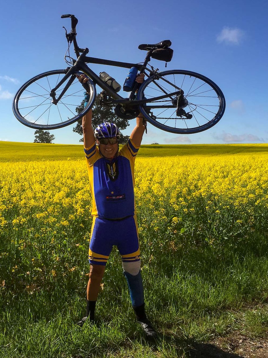 A cyclist with a prosthetic leg stands with a bike lifted above his head in a field of canola.