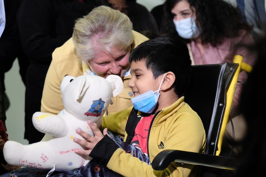 An indian boy in a yellow jacket smiling while being gifted a teddy from an elderly lady