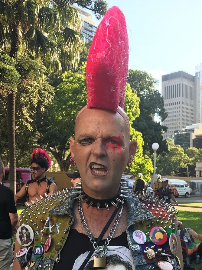 A man with a hot pink mohawk and studded denim vest and dog collar pulls a face to the camera.