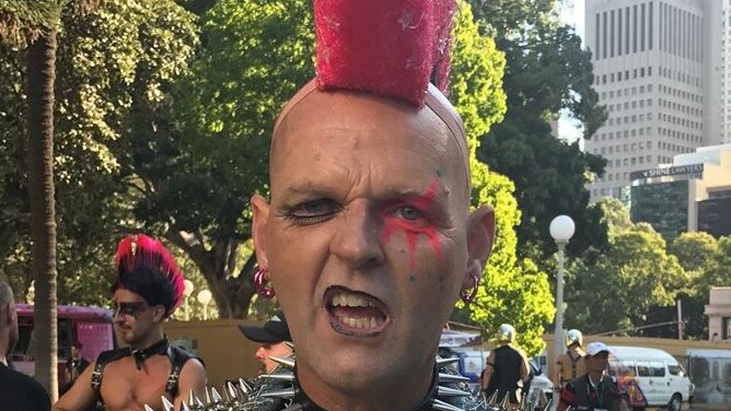 A man with a hot pink mohawk and studded denim vest and dog collar pulls a face to the camera.