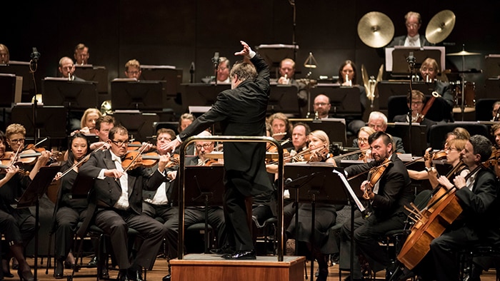 The Melbourne Symphony Orchestra on stage in performance. A conductor stands on the podium with his right arm raised.