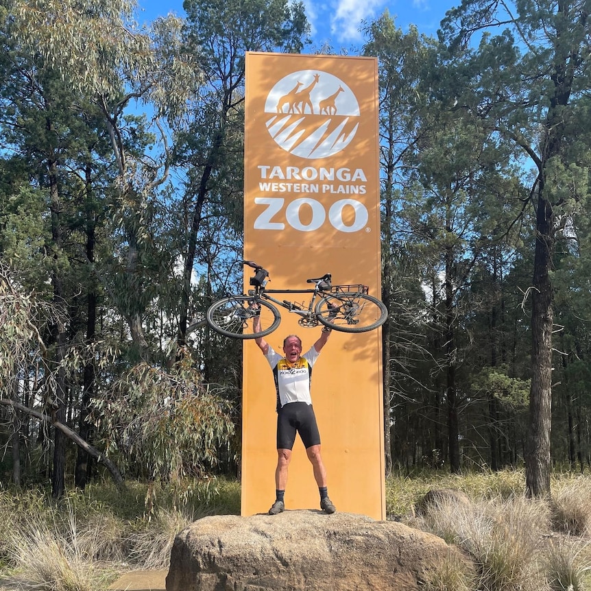 A smiling, older man holds a bike above his head in front of a large sign that reads "Taronga Western Plains Zoo".