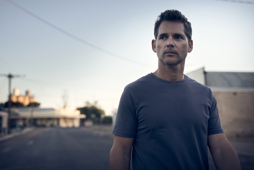 50-year-old white man with short salt-and-pepper hair wearing a blue tee shirt, with twilight country town streetscape behind.