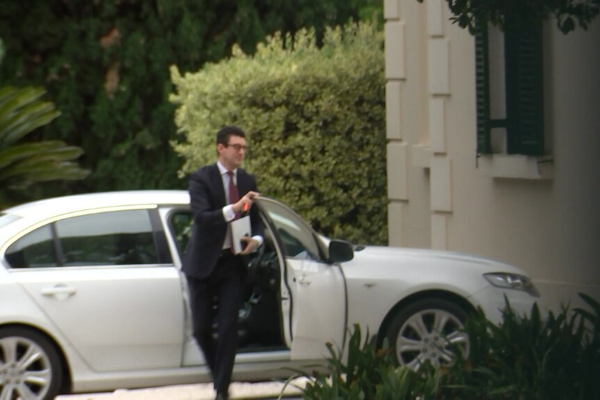 A man with dark hair, wearing a suit, steps out of a white sedan while holding a collection of papers