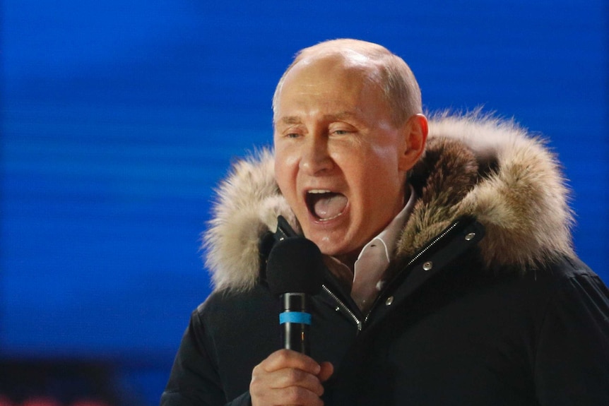 Vladimir Putin with his mouth open speaking into a microphone on stage.