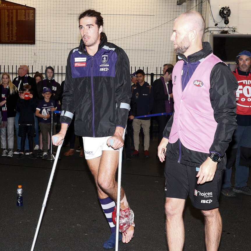 Dockers player Alex Pearce walks on crutches next to a team official in a pink vest inside the change rooms.