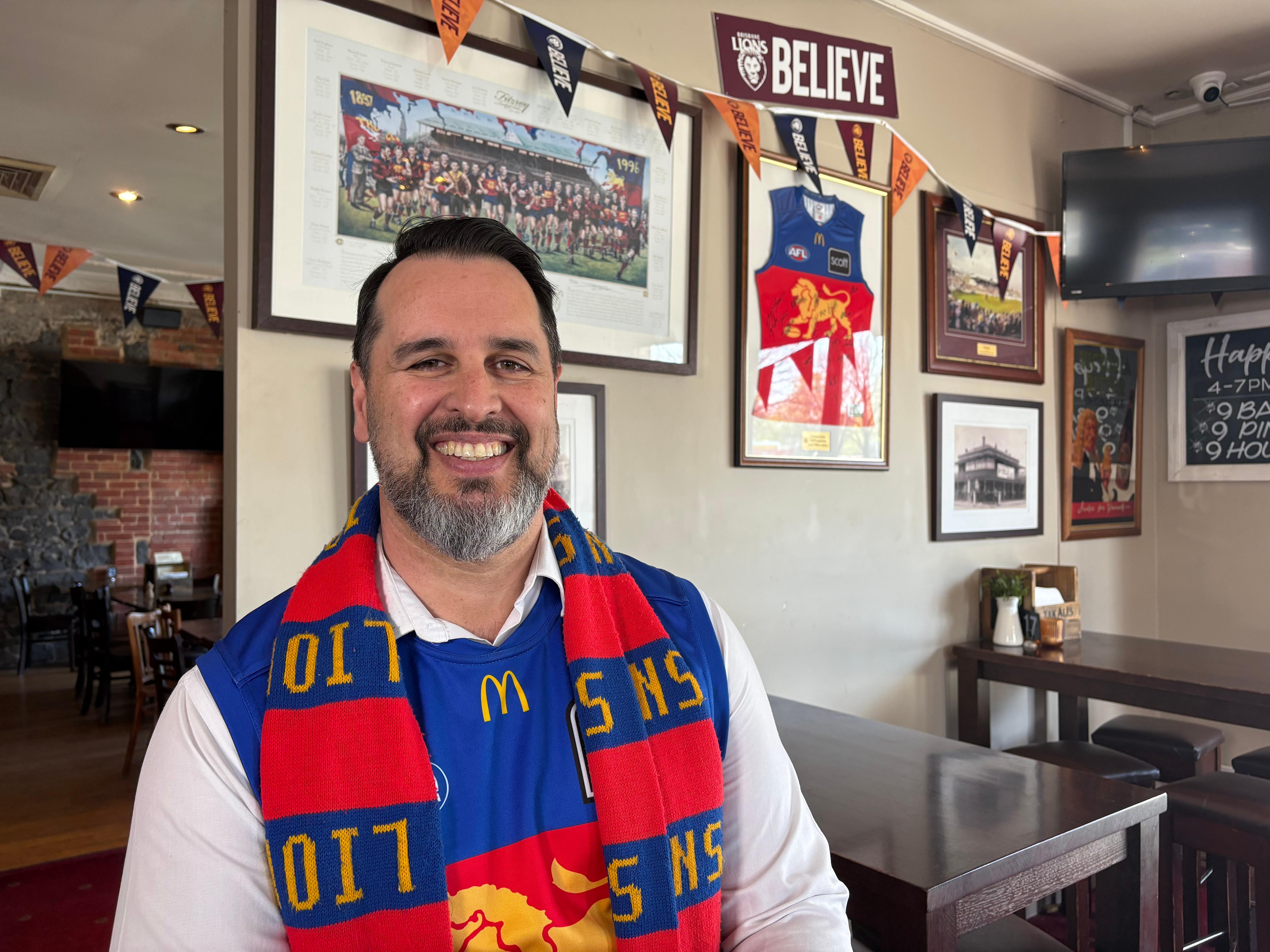Man smiling in front of Brisbane memorabilia wearing a Brisbane Lions jersey and scarf.
