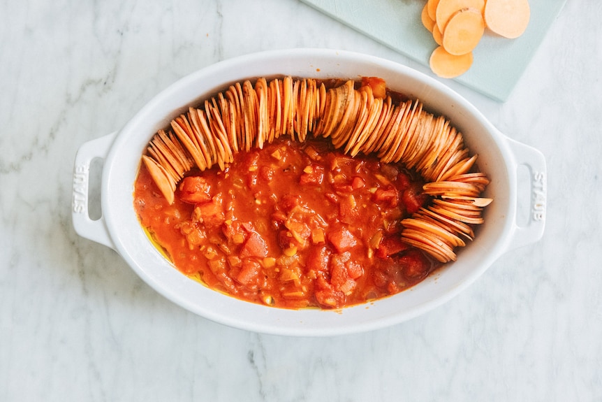 Bright red tomatoey sauce fills the base of a baking dish in which some thin slices of sweet potato have been arranged.