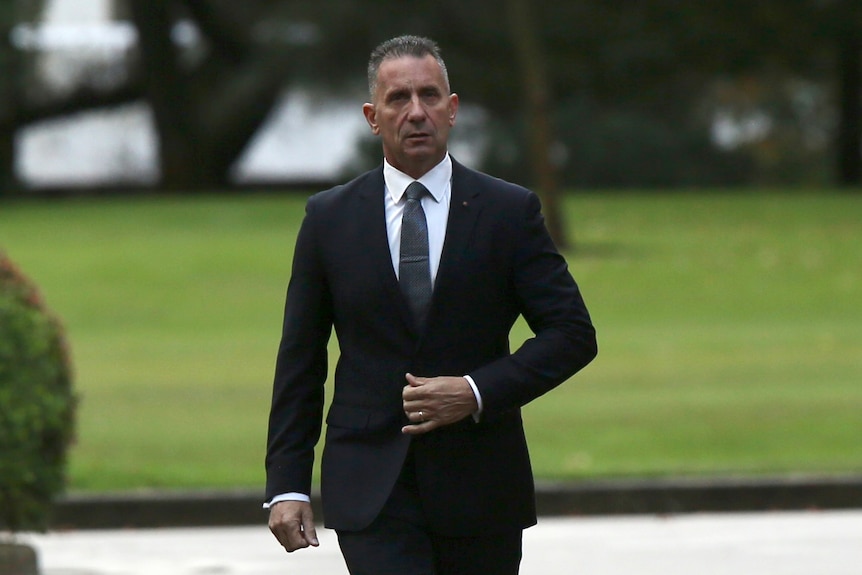 A mid-shot of Paul Papalia walking towards the camera wearing a dark suit and tie, with his left hand grasping his jacket.