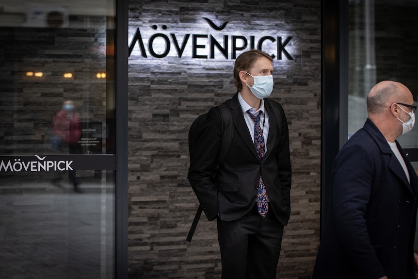 A man wearing a suit and a face mask walks away from a hotel.