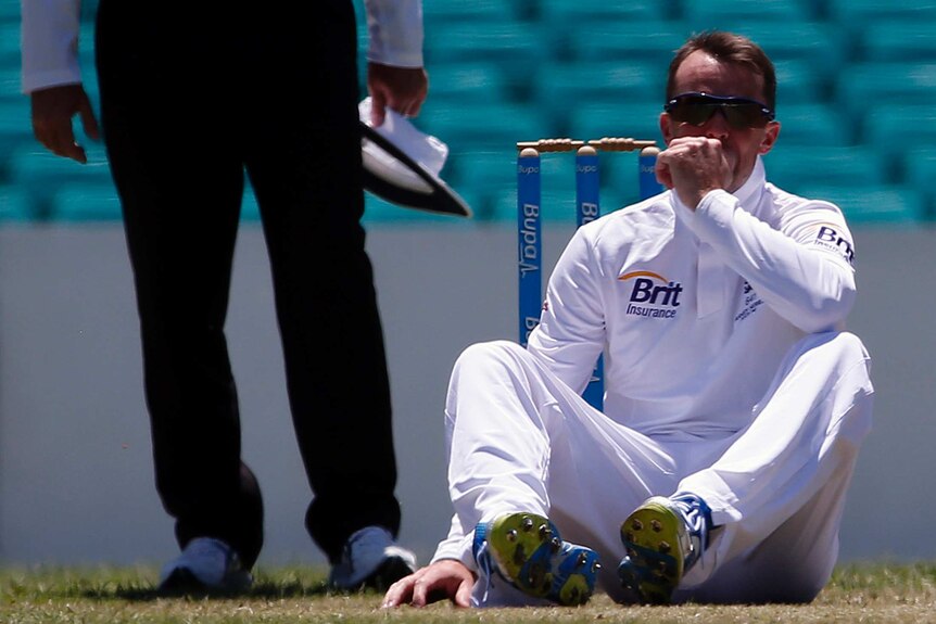 Cricketer in white sits on the pitch with stumps and an umpire in the background.