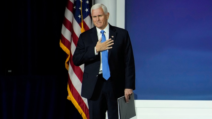 a man in a black suit, blue tie in front of an american flag 
