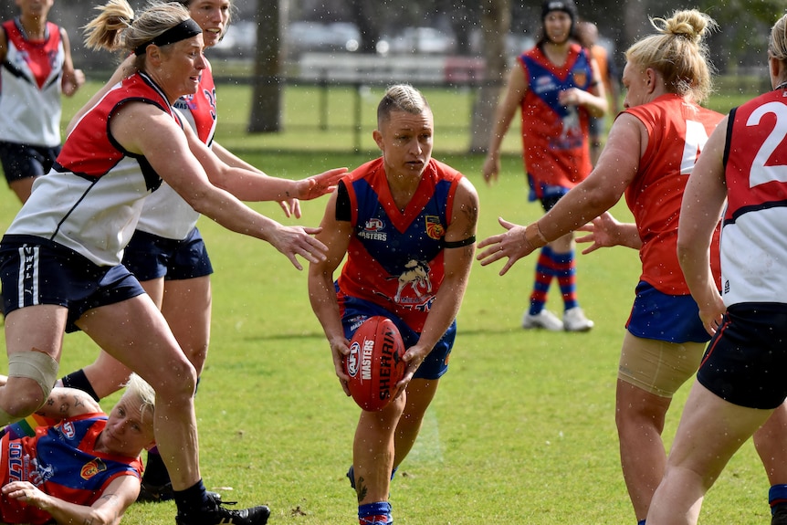 AFL masters player Millie Brown holds the ball and is running through players on either side. 