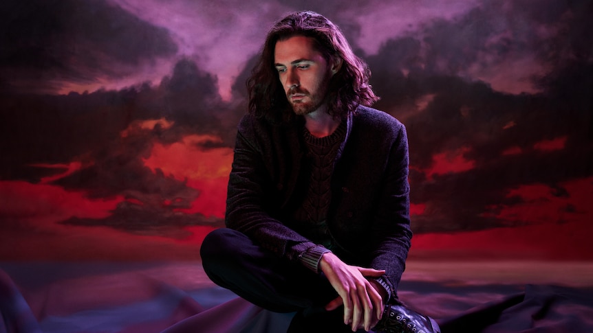 Hozier, a man with long dark hair, looks at the ground with despondent expression. A sunset is behind him.