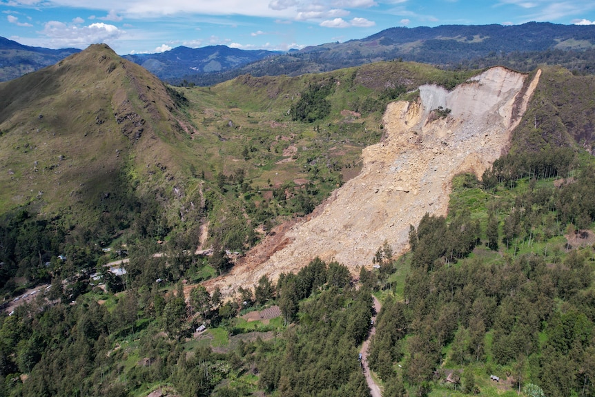 A large sandstone landslide is seen on the side of a sharp, green mountain range against a blue sky