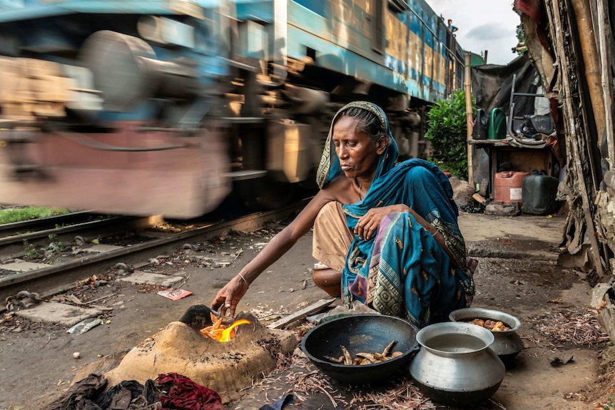 Bangladeshi woman wearing sari crouching by a fire and bowls of food next to a fast-moving train