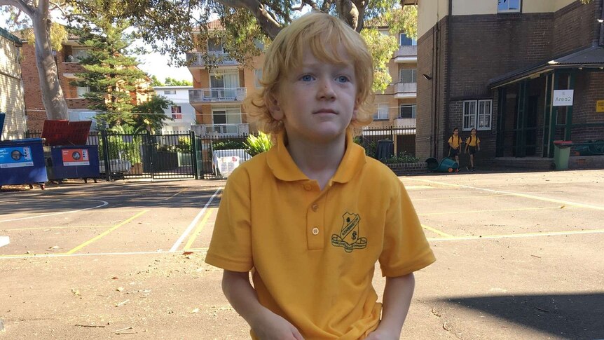 A boy with strawberry blond hair and a yellow school uniform sits in his school playground
