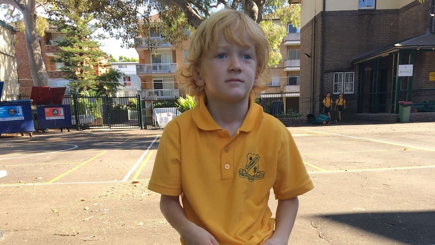 A boy with strawberry blond hair and a yellow school uniform sits in his school playground