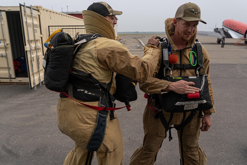 Two men in parachuting firefighting gear stand on an airplane tarmac. One checks the other's equipment.