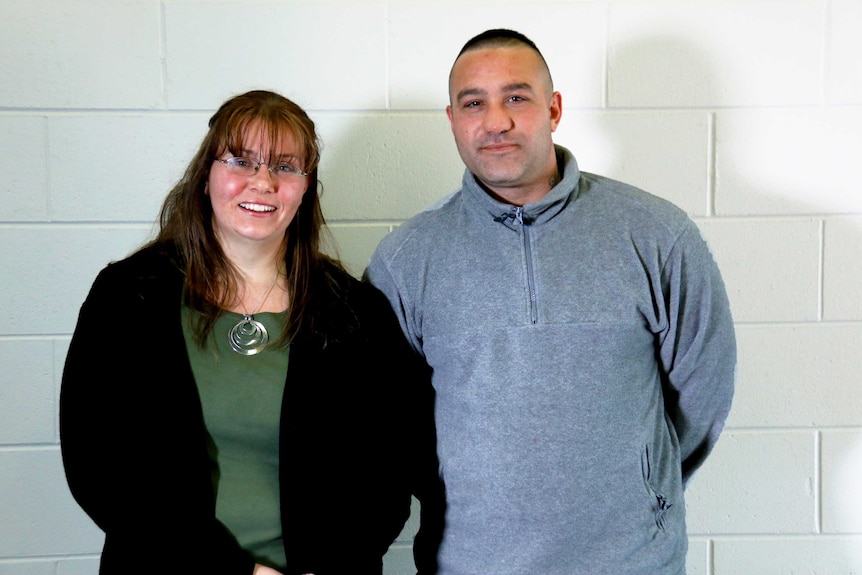 A woman and man stand next to each other, smiling, in front of a while brick wall.