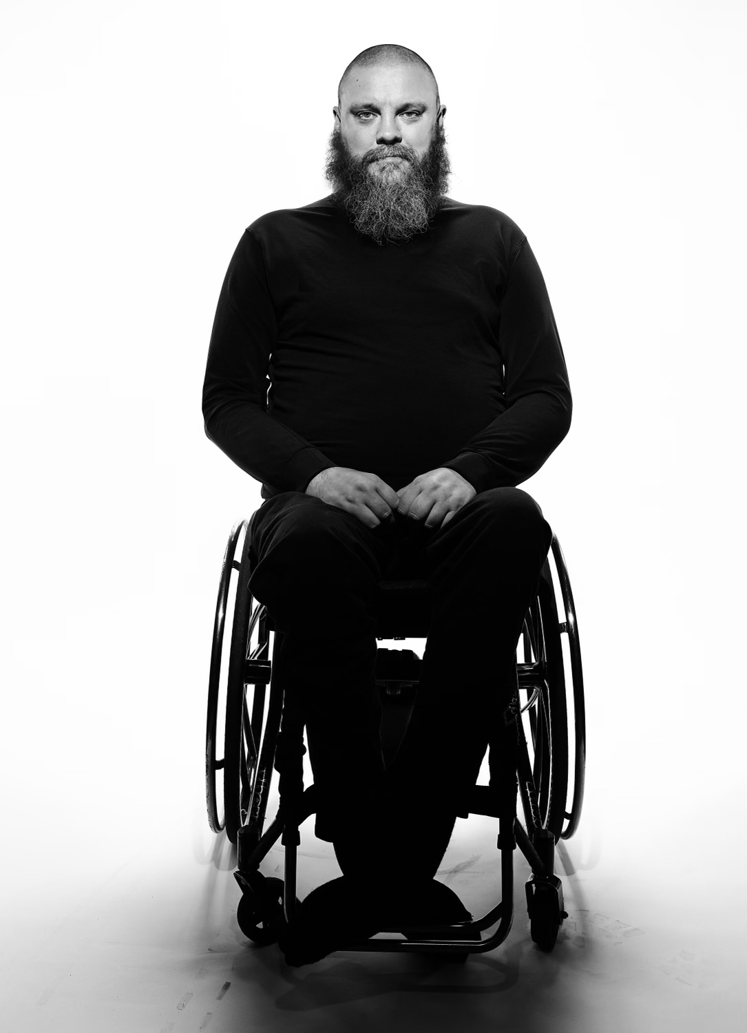 A black-and-white full-length portrait of a bald man with a long beard, dressed in dark clothing, sitting in a wheelchair.