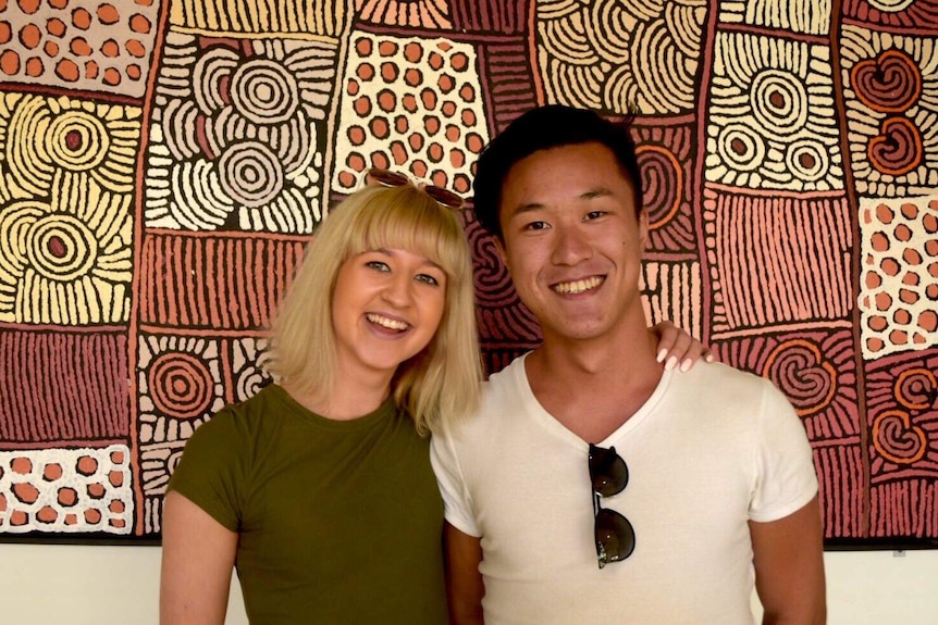A blonde Swedish woman and a man of Asian appearance from Hong Kong stand in front of a piece of Aboriginal art