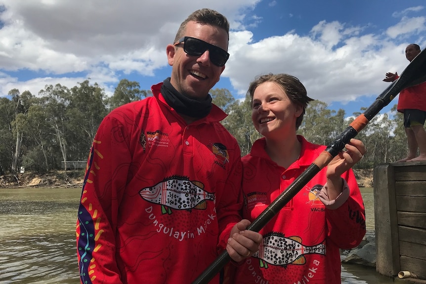 Man wearing red shirt and sunglasses stands next to a teenage girl holding a paddle in front of a river