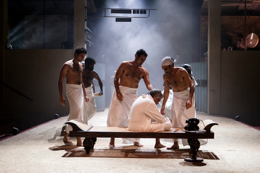 Men white in sarongs comfort a man in white kurta sitting with his head bowed in pain on a bench onstage.