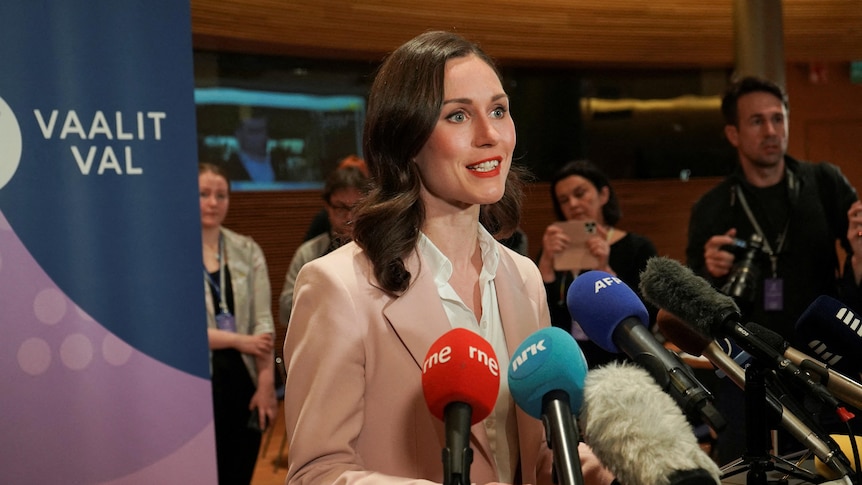 A white woman in her 30s, with medium-length brown hair &blue eyes. She's at a media conference with microphones pointed at her.