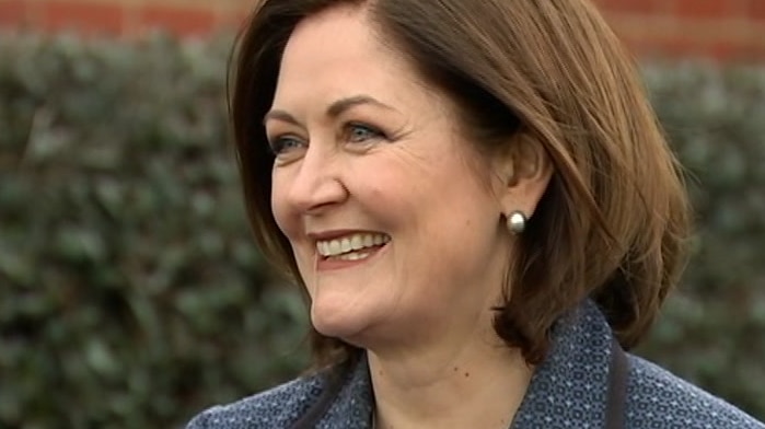 Sarah Henderson grins as she stands in front of a brick wall in a suit giving a press conference.