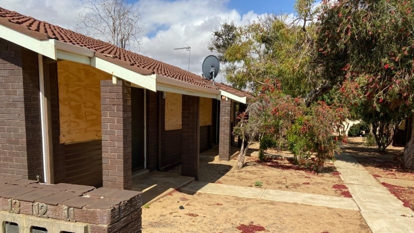 A brown brick house has boarded up windows and dry grass.