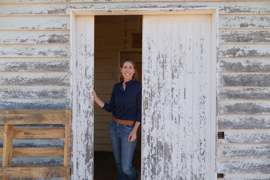 A smiling woman wearing blue shirt, jeans, tan belt, stands in a doorway of an old building with peeling paint.