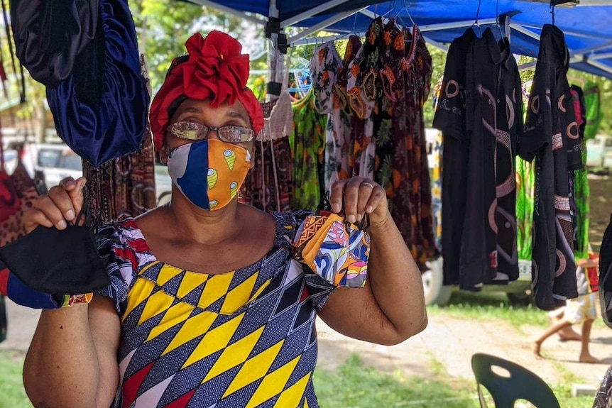 A middle-aged black woman with red hairnet & colourful dress holds up two colourful masks in market.